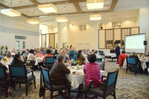 Residents seated at round tables attending a presentation in a large community dining room.